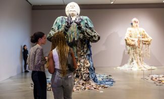 Two people stand in an art gallery in front of large, figurative sculptures with textured, layered draped outfits