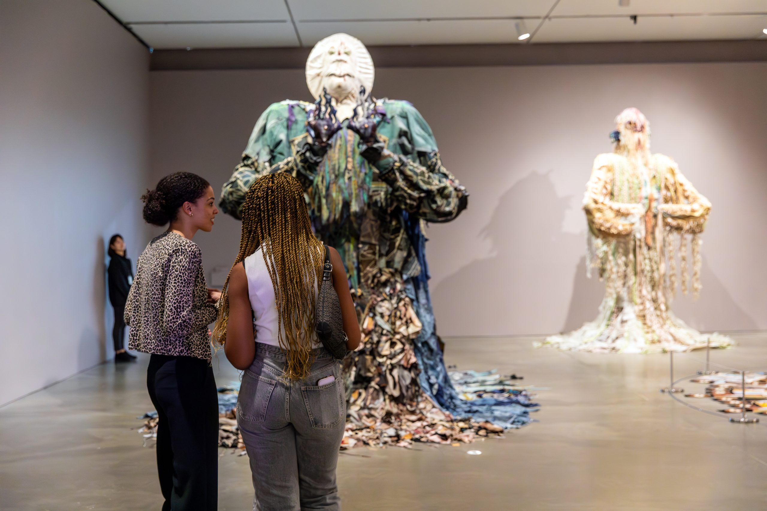 Two people stand in an art gallery in front of large, figurative sculptures with textured, layered draped outfits