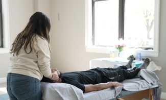 A woman with long dark hair touches the face of a young man lying on a massage table in a brightly lit room.