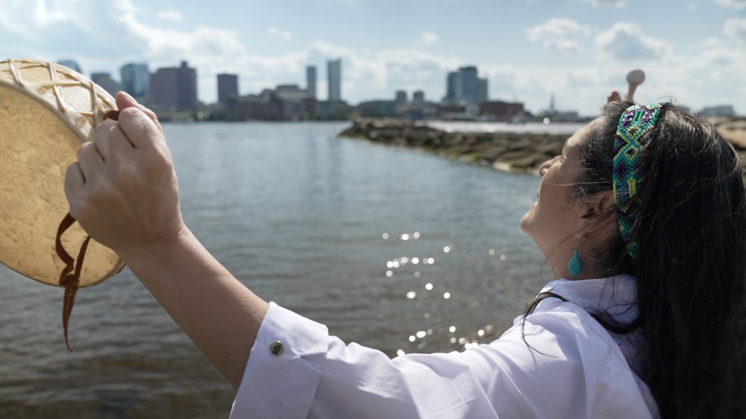 A woman with dark hair and a white blouse stands by the water, holding a small drum, with the Boston skyline in the distance.