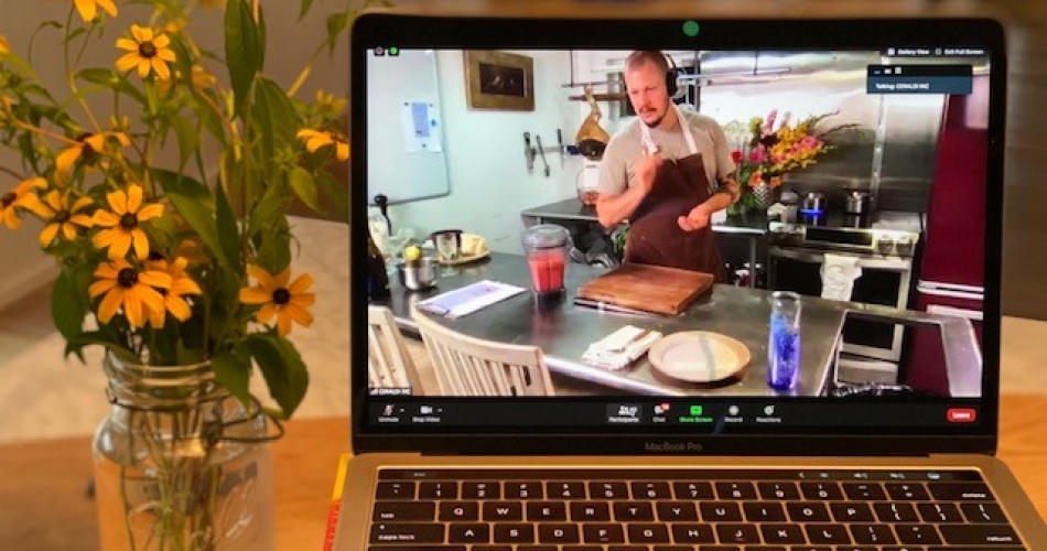 A laptop showing a chef in a profession kitchen sits next to a jar of cut flowers.