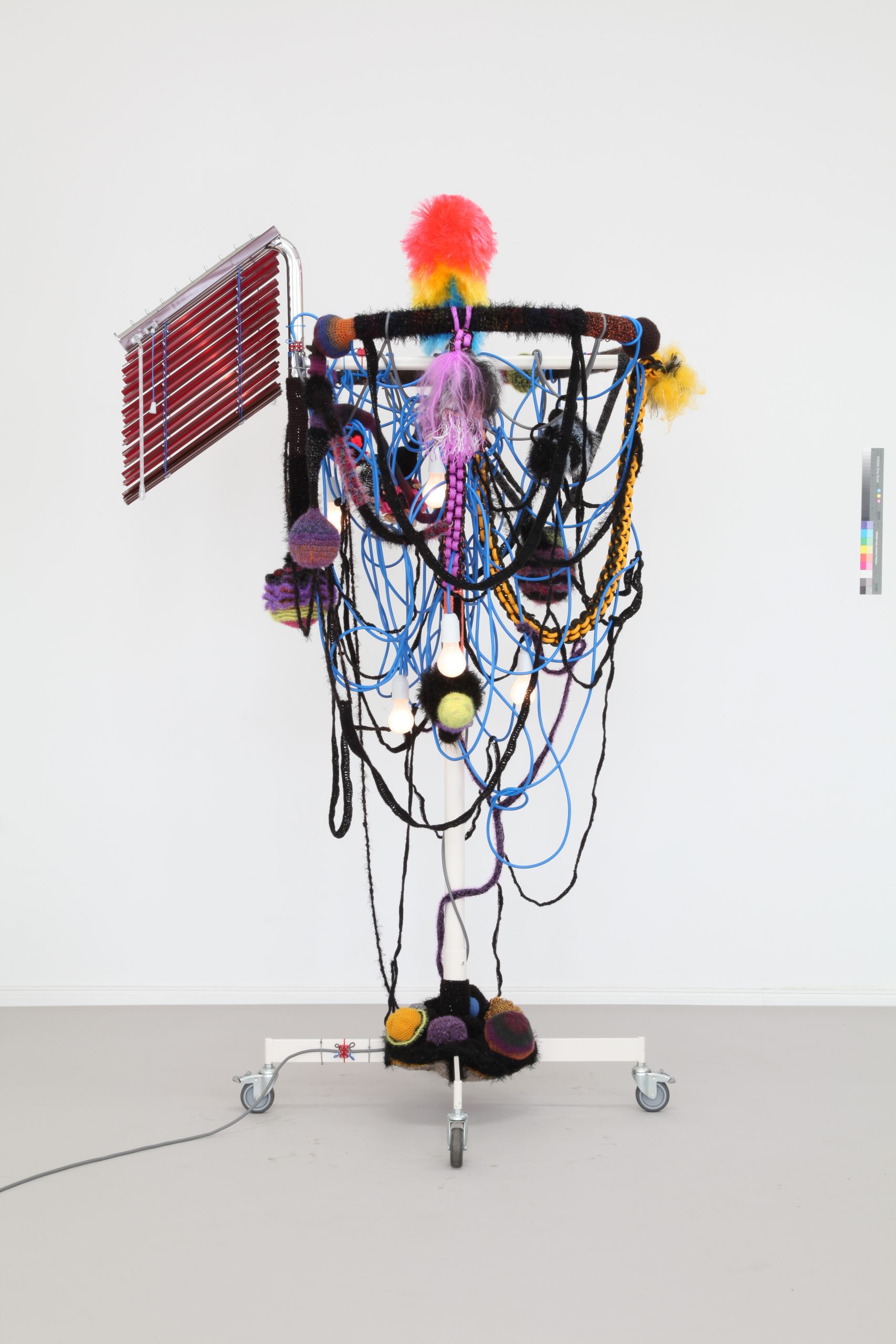 Photograph of sculpture made from a clothing rack draped in cable, rope, cord, light bulbs and aluminum blinds.