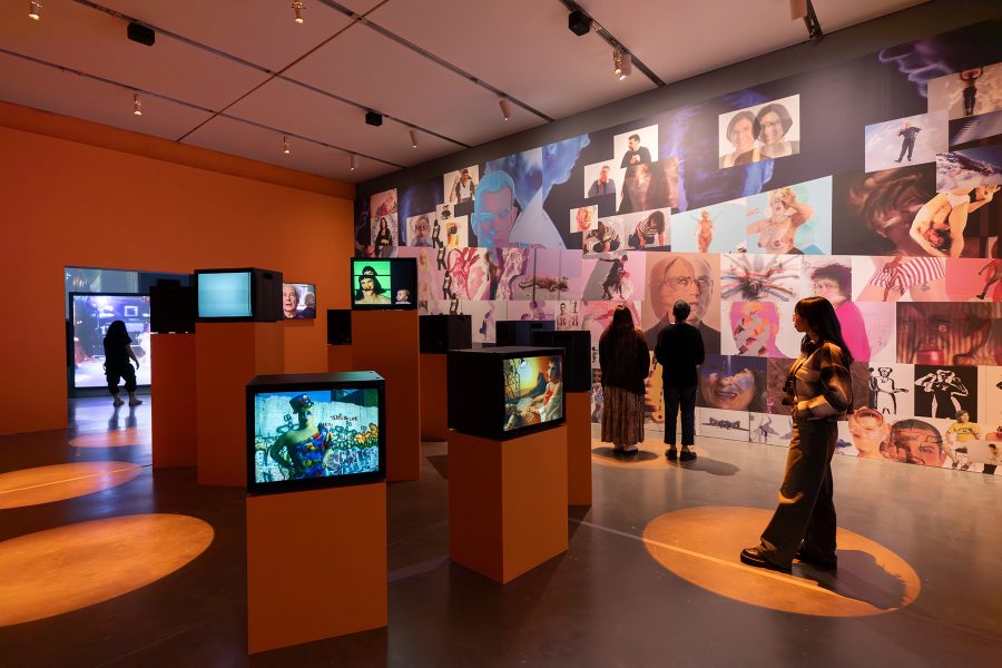 Orange gallery with small TV monitors on pedestals arranged in spiral and a collaged back wall