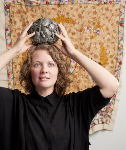 Person with shoulder length hair holding metal ball on their head in front of a quilted work on the wall