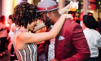 At ICA First Fridays in March, a couple dances closely at the party. The woman with curly hair wears a patterned dress, while the man with glasses dons a hat and red suit. He holds a drink as they embrace, surrounded by a lively crowd.