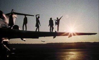 Five people stand on the wing of an airplane with a sunset in the background