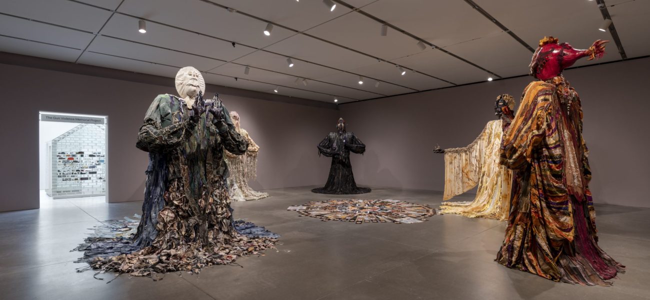 Large figurative sculptures draped in patterned, flowy fabrics in a darkened gallery with a white house structure in view in the doorway of the next gallery