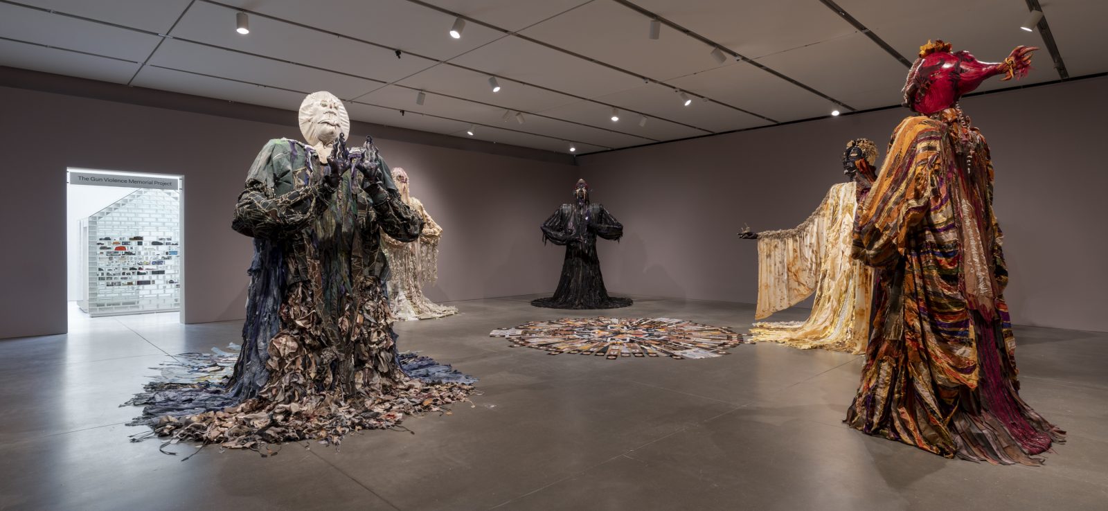 Large figurative sculptures draped in patterned, flowy fabrics in a darkened gallery with a white house structure in view in the doorway of the next gallery