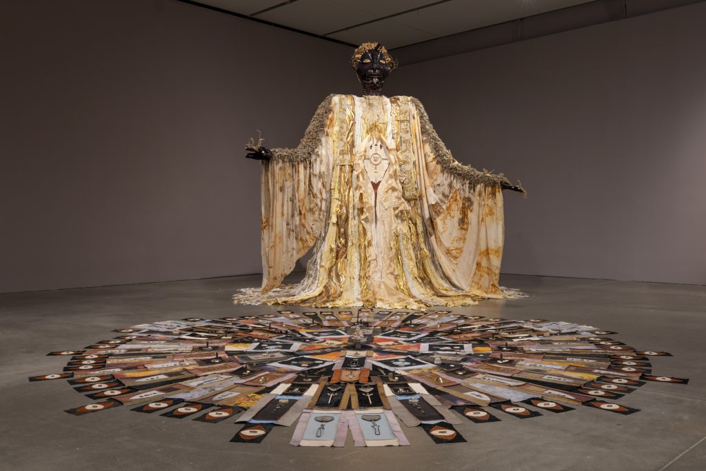 A tall figurative sculpture draped in a gold robe with a circular patterned floor work in front of it