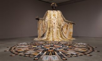 A tall figurative sculpture draped in a gold robe with a circular patterned floor work in front of it