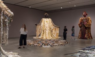 Visitors in a darkened gallery looking at large figurative sculptures draped in patterned fabrics