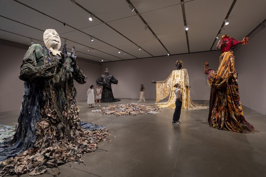 Visitors in a darkened gallery looking at large figurative sculptures draped in patterned fabrics