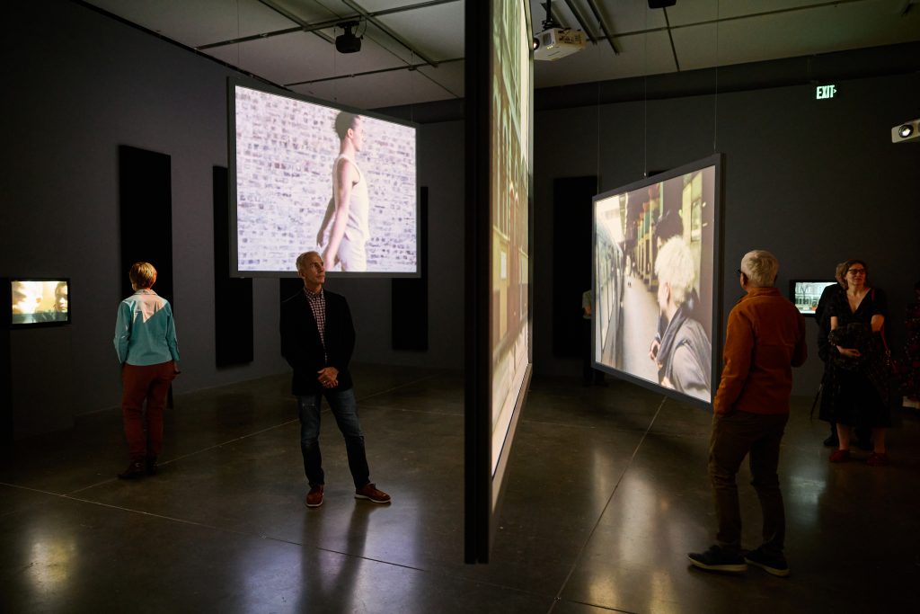 Visitors in a darkened gallery watching large, suspended video screens hanging at different angles and heights