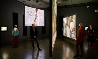 Visitors in a darkened gallery watching large, suspended video screens hanging at different angles and heights