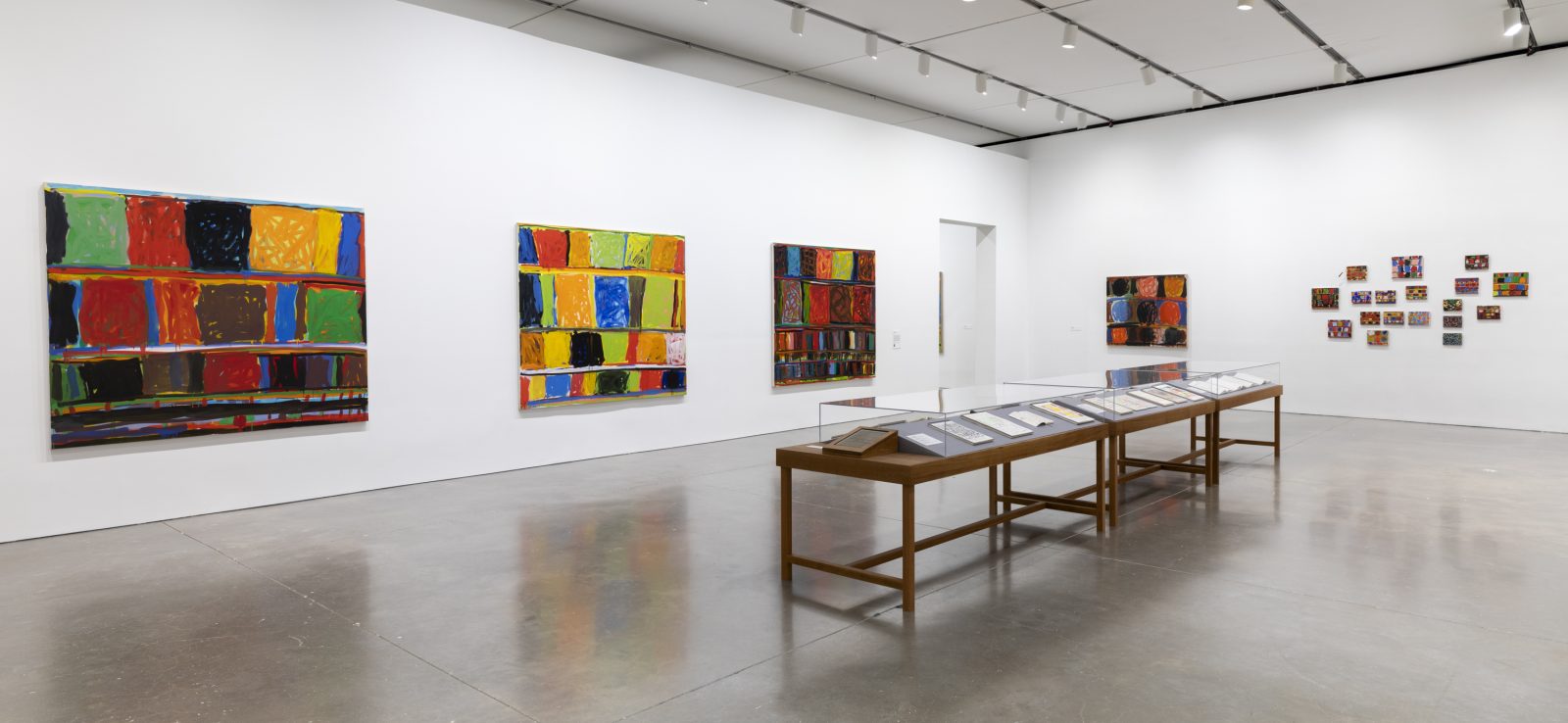 A gallery with white walls at ICA Boston displays colorful abstract paintings with a series of small works clustered on one wall, and two wooden display tables with small works on paper under glass. The floor is polished concrete.
