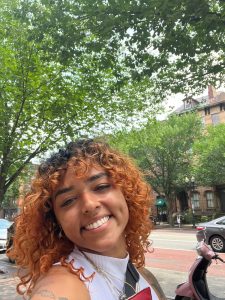 Selfie photo of person with copper, shoulder-length curly hair smiling outside