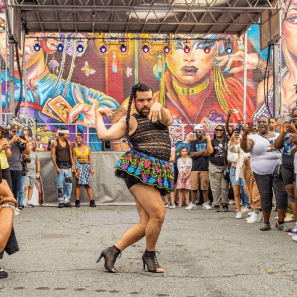 A person energetically dances in a colorful outdoor setting, wearing a black lace top and floral skirt, surrounded by vibrant murals. The engaged audience watches intently, capturing photos of the lively performance.