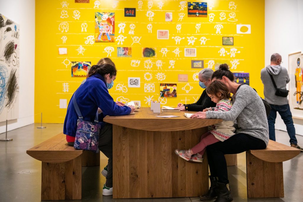 People sitting at a wooden table in an art gallery, engaging in drawing activities. The wall behind them is bright yellow and decorated with various colorful artworks and abstract designs.