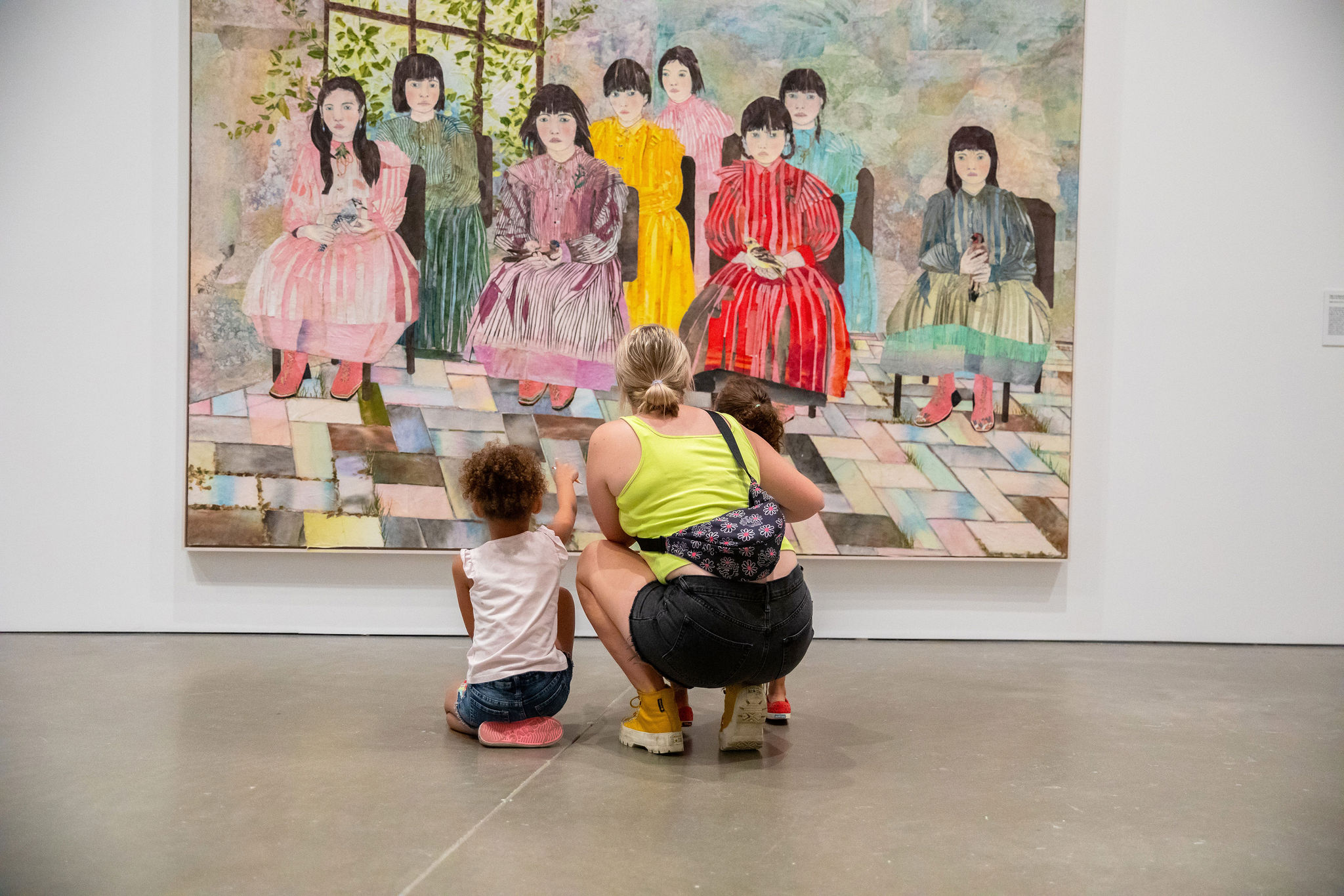 An adult crouches to look at a large painting with two children