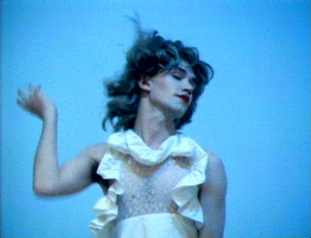 A video still showing a fair-skinned person in a white dress and red lipstick in motion.