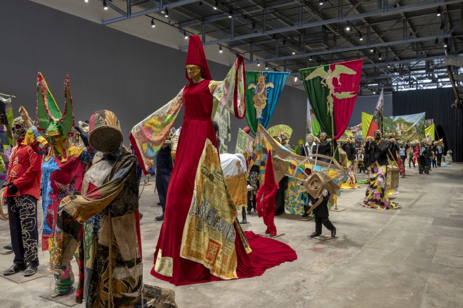 A mass of figurative masked sculptures march forward with a tall red hooded figure near the front