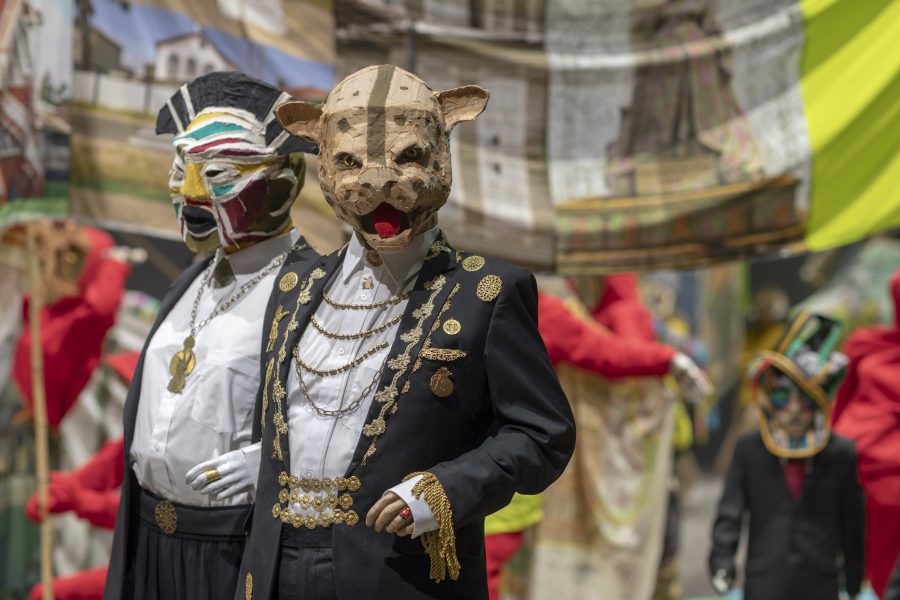 Two masked sculptures in matching black suits with gold adornments in front of a large banner