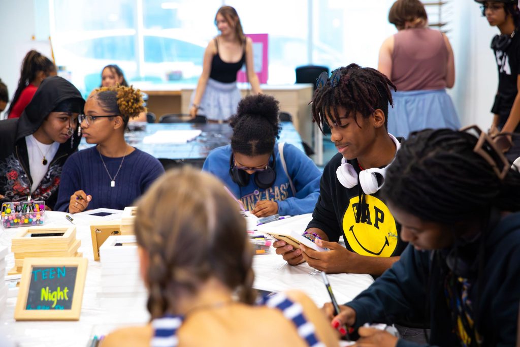 A group of teens engaged in activities at a table. One wears a black shirt with a yellow smiley face with a word instead of eyes and headphones. Art supplies and small boxes are scattered around as others chat or focus on their projects in a bright, casual setting dedicated to arts education.