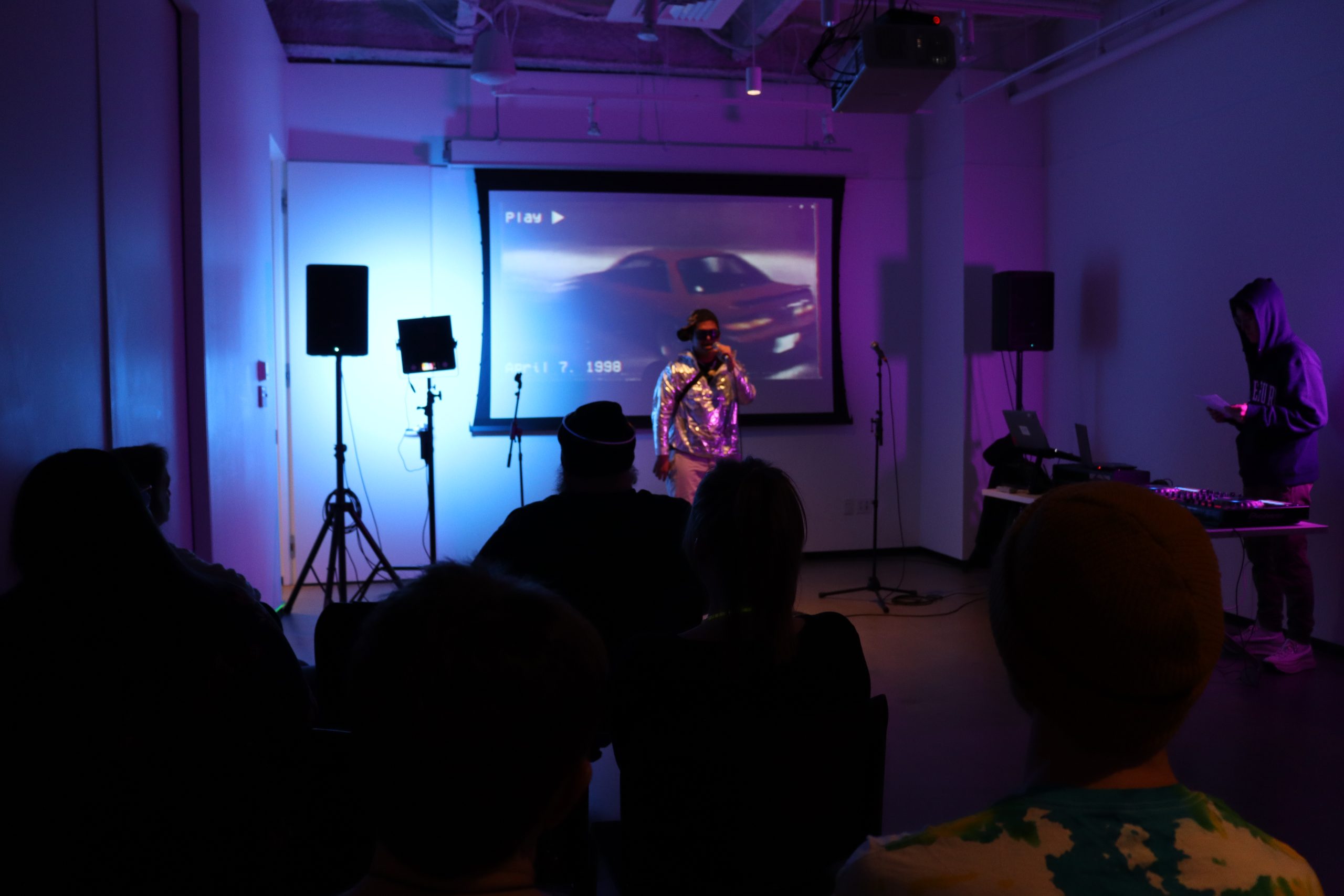 A person performs on stage in a dimly lit room, with an audience seated in silhouettes. A projector displays a car on a screen. Equipment and a DJ are set up nearby, and the lighting casts a blue-purple hue.