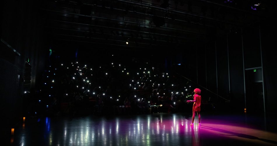 A performer stands on a dimly lit stage facing an audience. The audience is mainly in darkness, with multiple small lights like phone screens illuminating the seating area, creating a starry effect. The stage has a purple spotlight on the performer.