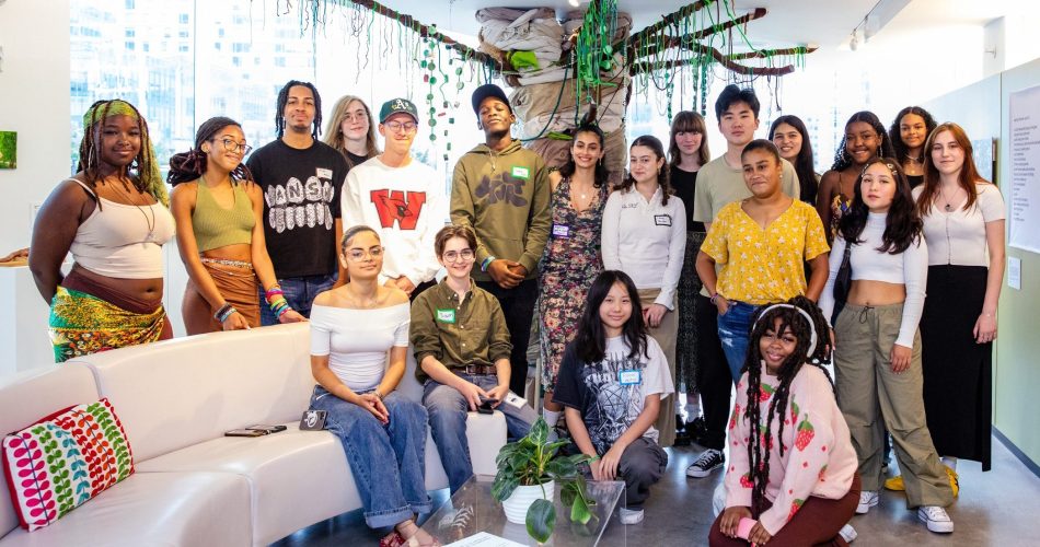 A diverse group of people standing and sitting in a well-lit room with colorful decor. There are various green plants hanging from the ceiling. Everyone is smiling and looking at the camera. Tables and informational posters are visible in the background.