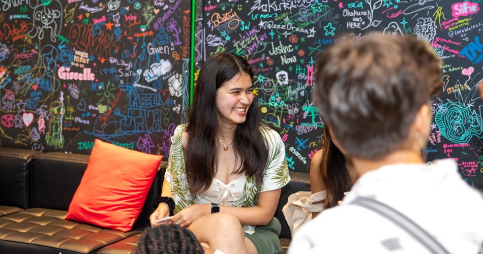 A woman with long hair sits on a black couch, smiling and engaging with people around her. Behind her is a colorful wall filled with graffiti and signatures. There are red cups on the table in front.