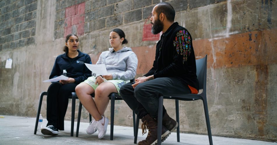 Three people sitting on chairs in a discussion against a brick wall. One person on the right has a colorful design on their black jacket. The person in the middle holds paper, and the person on the left gestures with their hand.