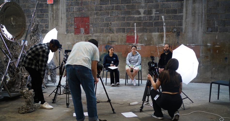 A group of people is filming in an industrial setting. Three individuals sit in chairs facing the cameras, with two people operating the cameras. Bright studio lights illuminate the scene. The background features a textured concrete wall.