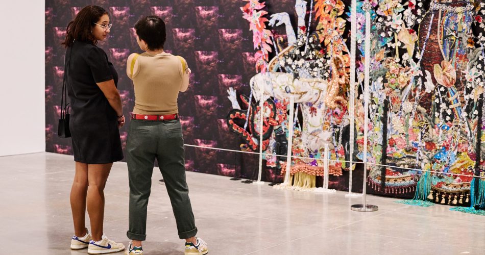 Two people stand in an art gallery, observing a large, colorful tapestry featuring intricate floral and abstract patterns. The gallery has polished concrete floors and white walls.