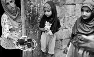 three people appear in a black and white photo. from left to right is a woman in a hijab offering tea to two girls. one girl in the middle eats bread and looks at the tea. the girl on the far right looks off into the distance.