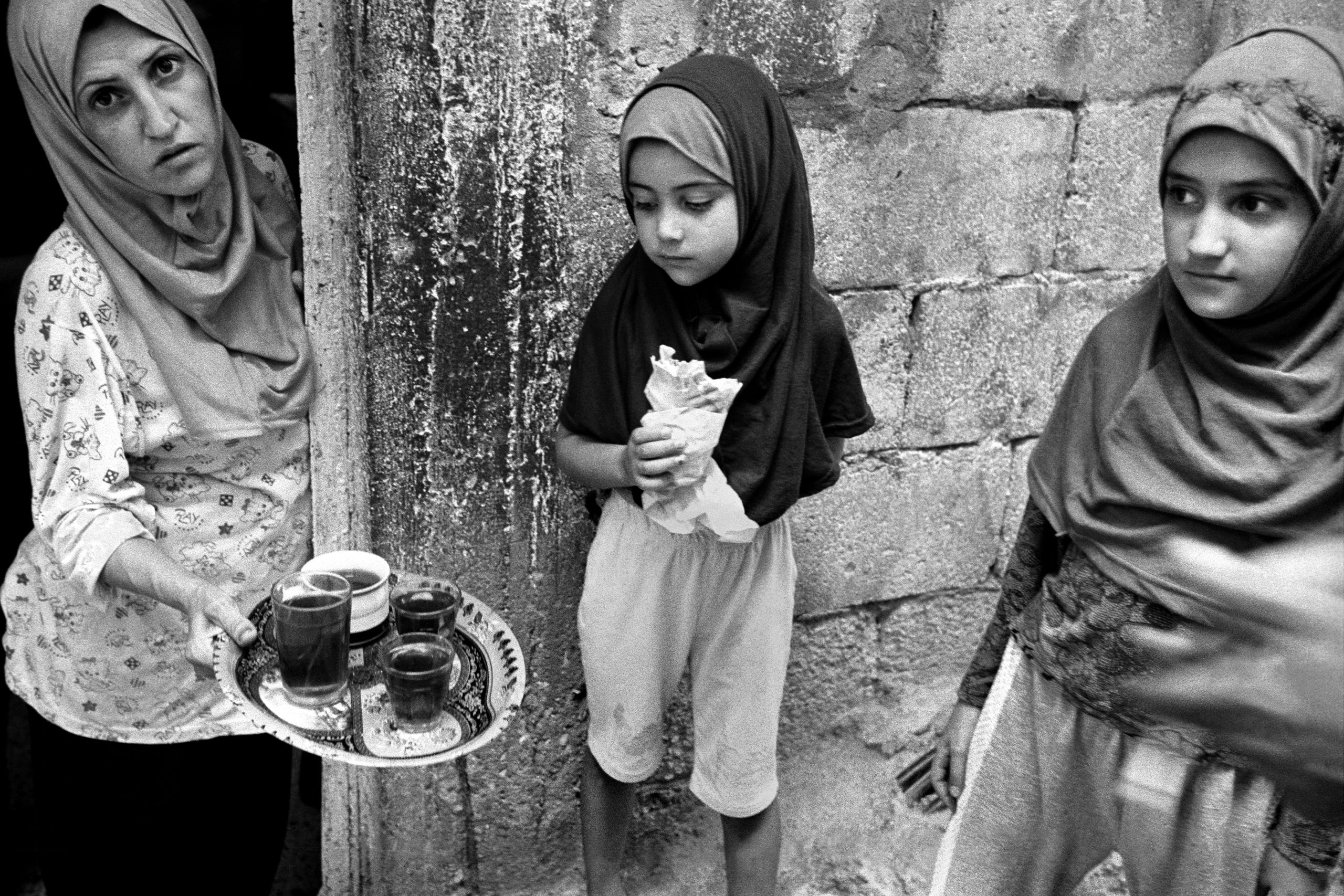 three people appear in a black and white photo. from left to right is a woman in a hijab offering tea to two girls. one girl in the middle eats bread and looks at the tea. the girl on the far right looks off into the distance.