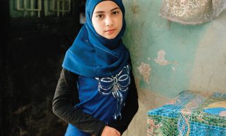 a girl in a blue Hijab stands in front of a domestic space. she wears a blue shirt with a silver bow. in the top right corner is gold and pink fabric in the shape of a bow.