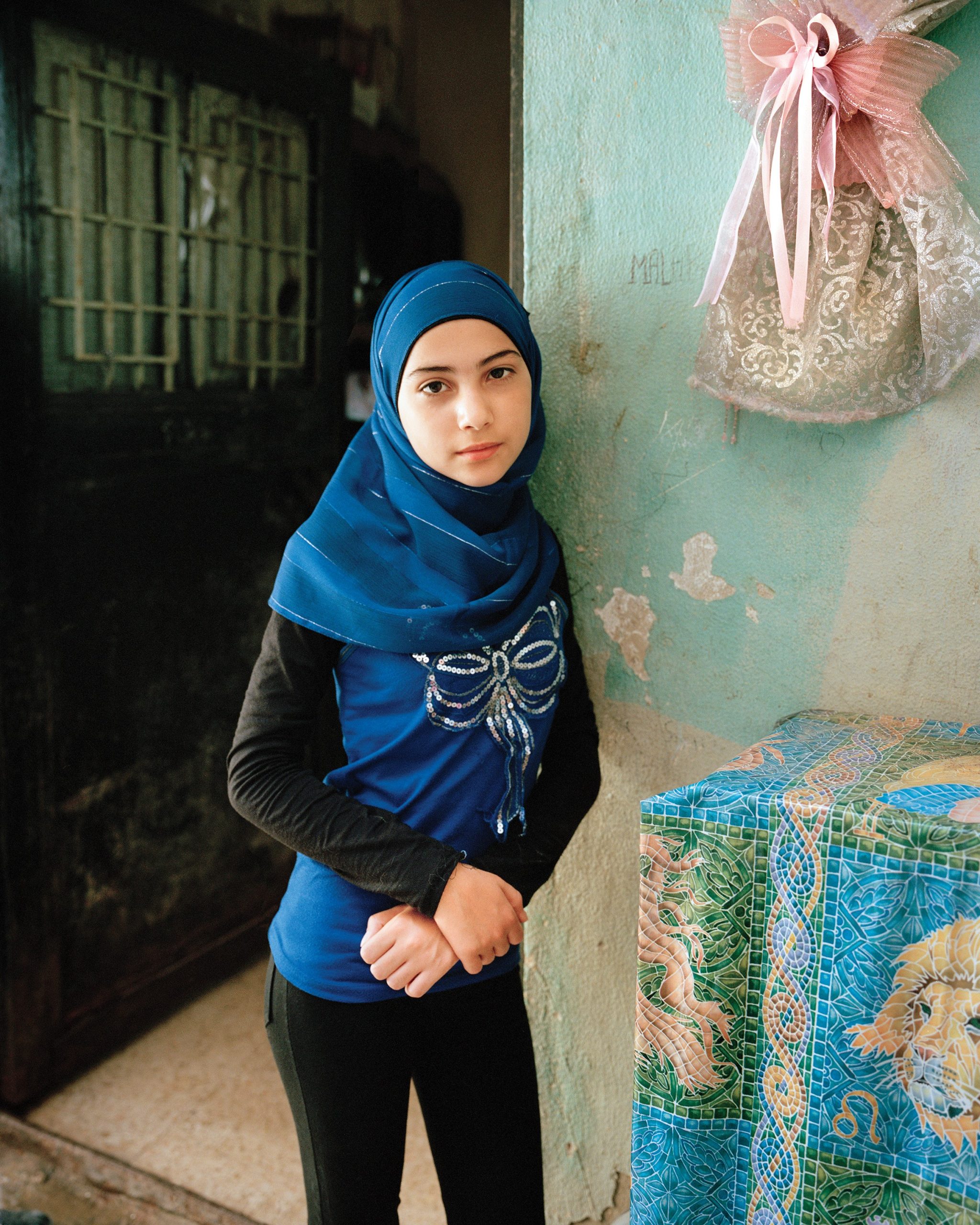 a girl in a blue Hijab stands in front of a domestic space. she wears a blue shirt with a silver bow. in the top right corner is gold and pink fabric in the shape of a bow.