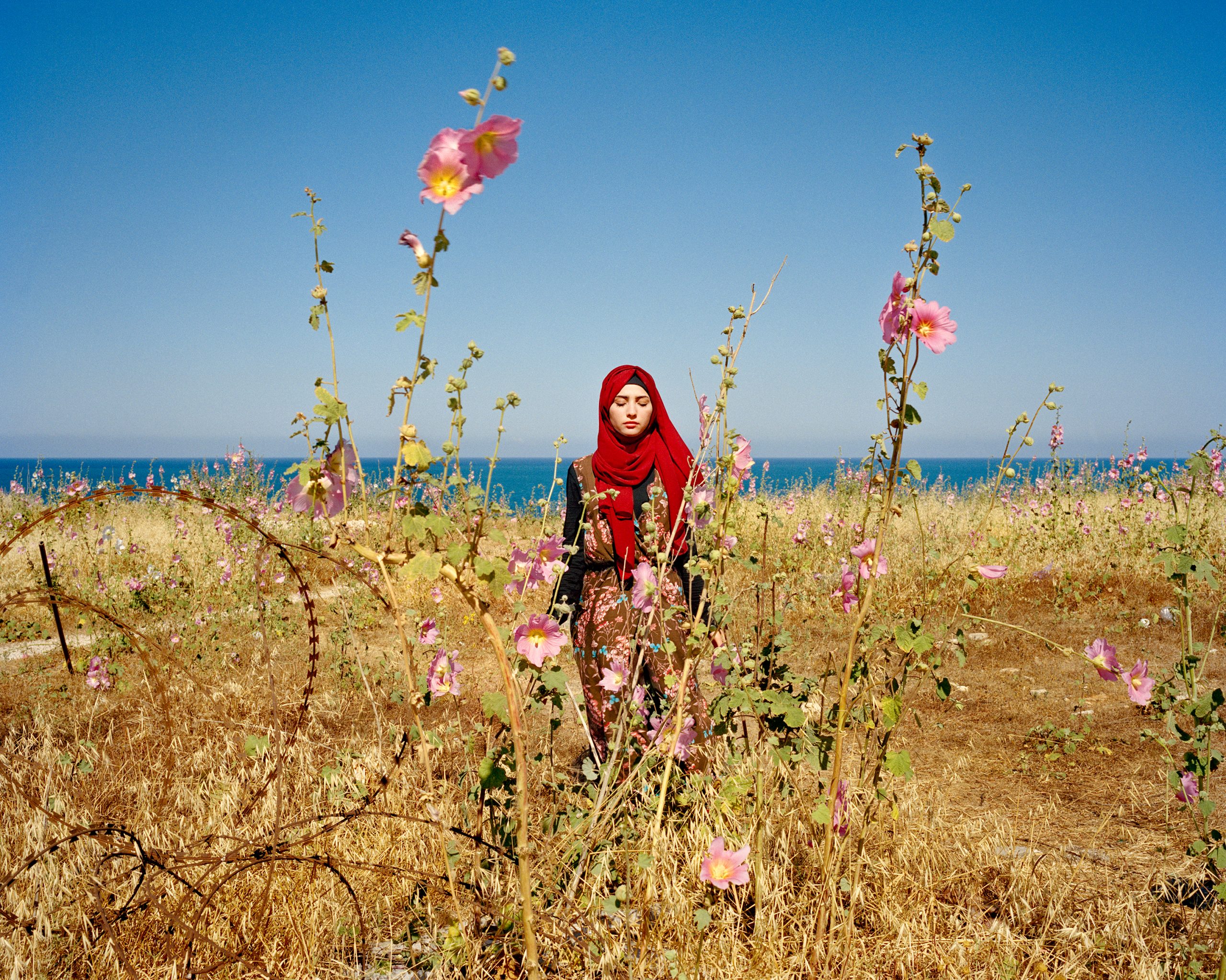 a girl in a red hijab stands in a field of pink flowers. her eyes are closed. behind her is the open ocean