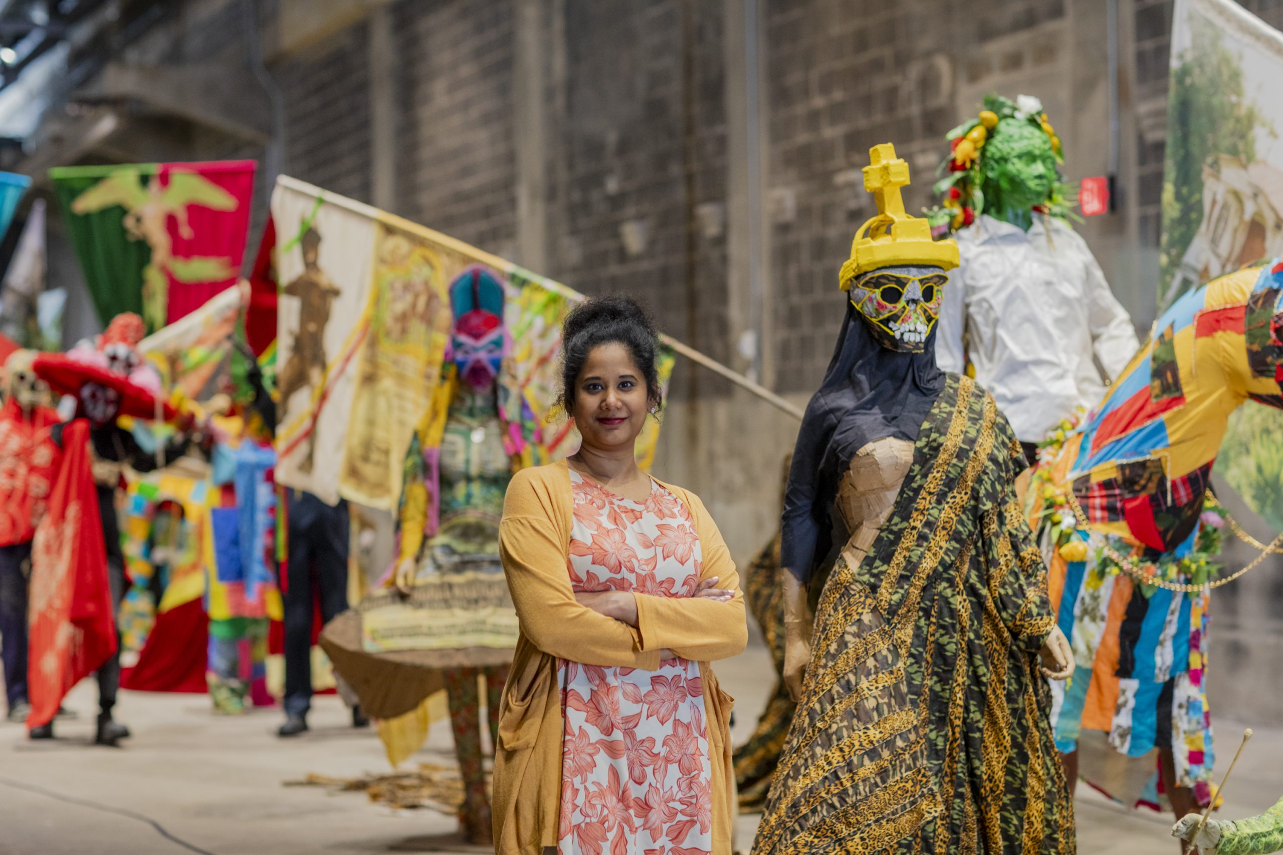 A person with medium dark skin and a black bun in front of colorful masked sculptures arranged in a procession in an industrial space
