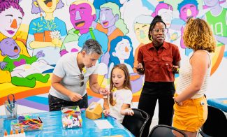 Four people stand and make crafts in front of a mural