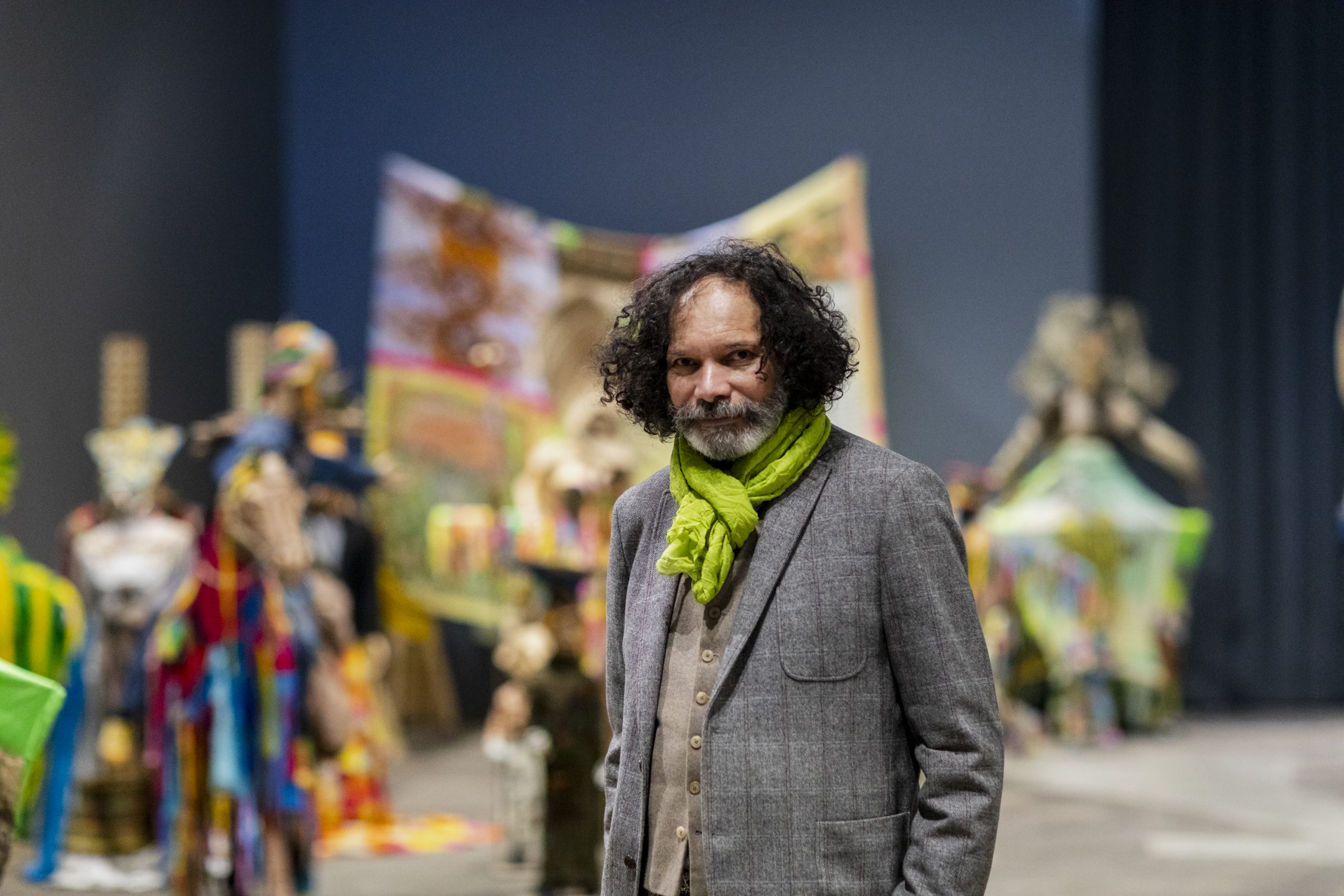 A older person with curly dark hair and white beard stands in front of sculptures out of focus