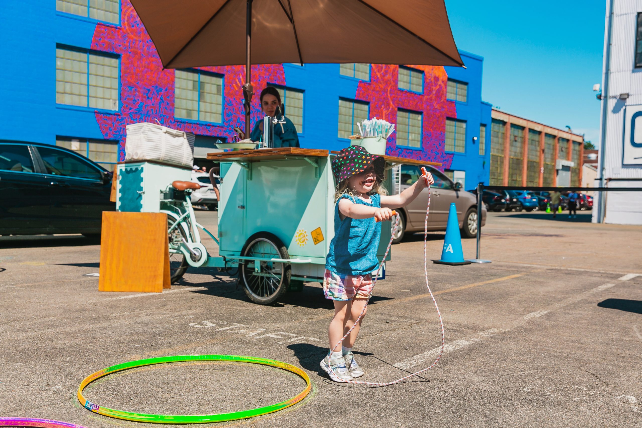 A young child in bright clothing plays with a hula hoop and jump rope outside in East Boston