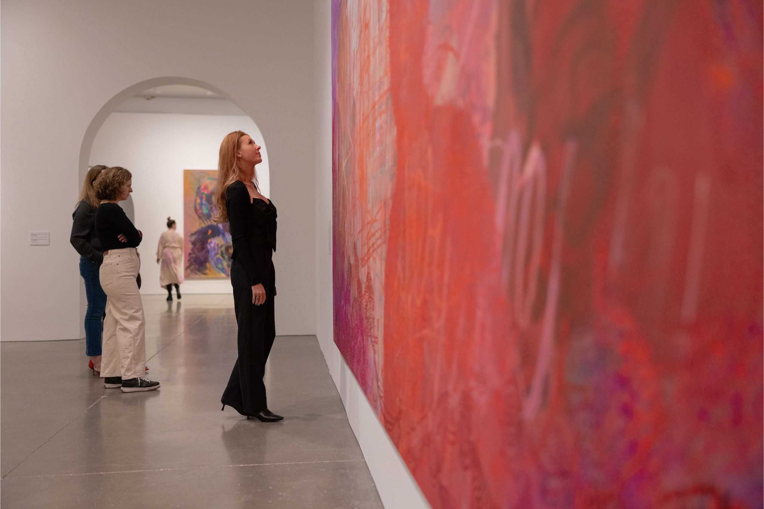 a woman in a black outfit looks up at a red and pink painting in a gallery space. behind her two women are in conversation