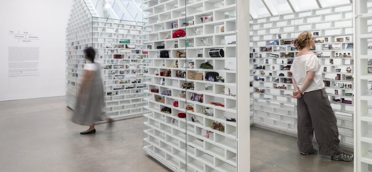 Two visitors look at white house shaped installations with objects slotted into brick-shaped shelves