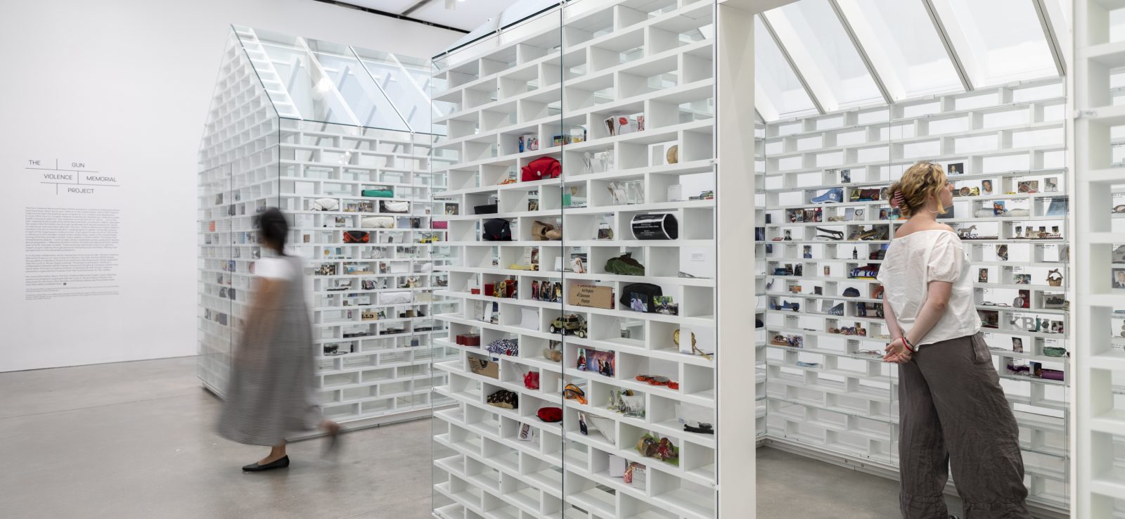 Two visitors look at white house shaped installations with objects slotted into brick-shaped shelves