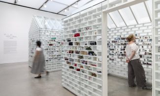 Two visitors look at white house shaped installations with objects slotted into brick-shaped shelves