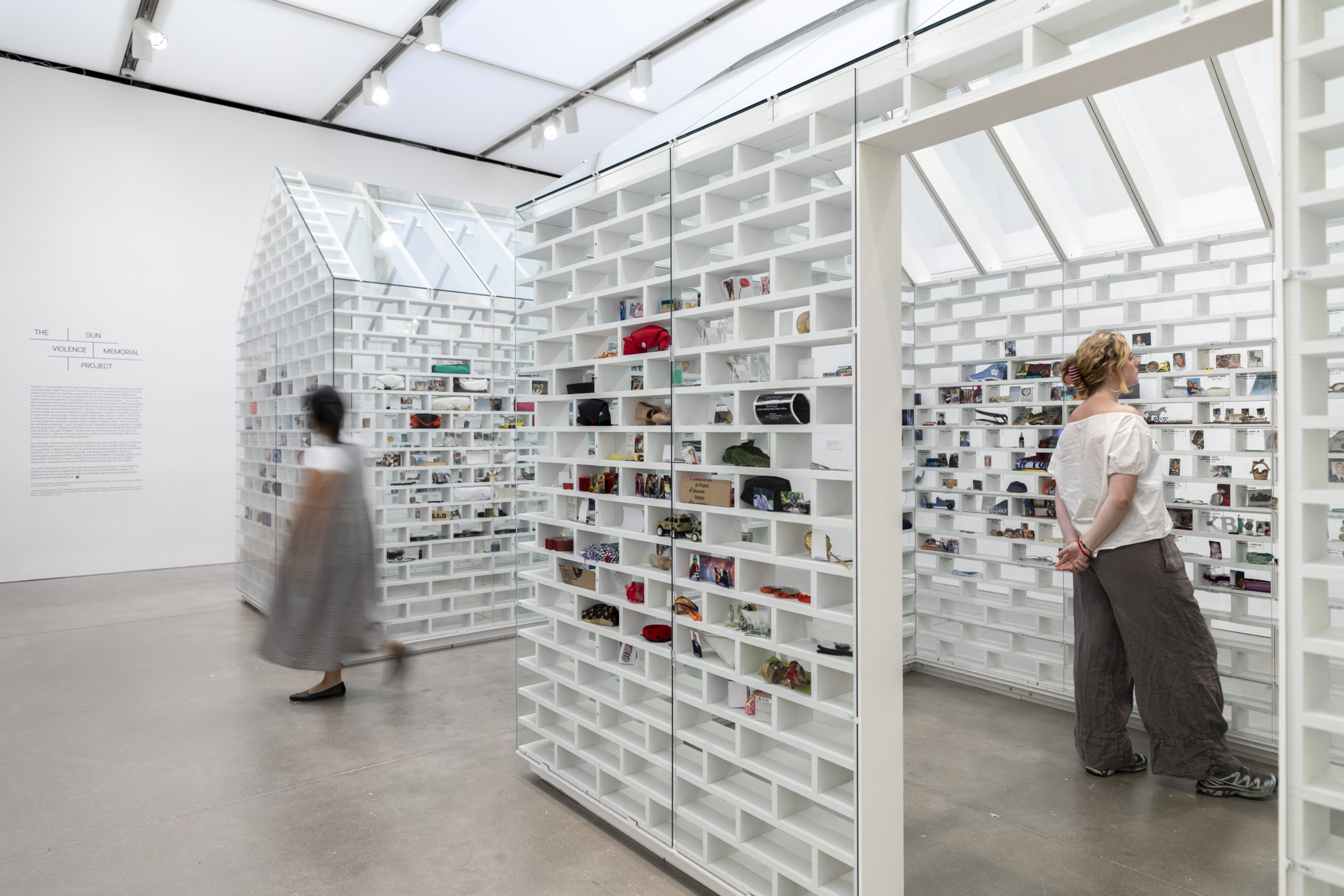 Two visitors look at white house shaped installations with objects slotted into brick-shaped shelves