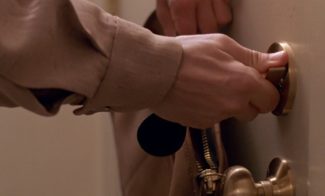 A film still of a hand in a beige sleeve turning a brass lock on a door.