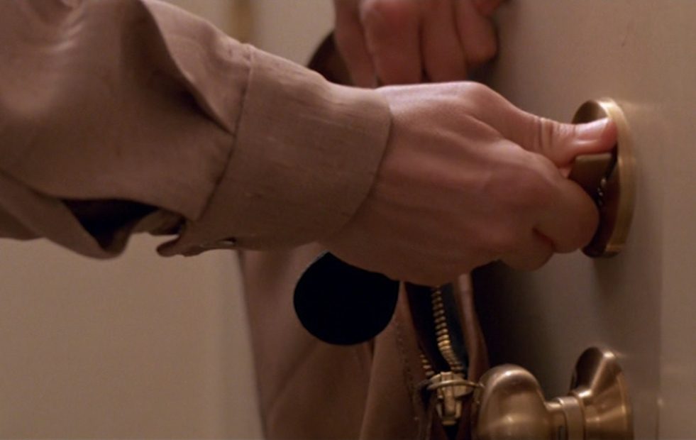 A film still of a hand in a beige sleeve turning a brass lock on a door.